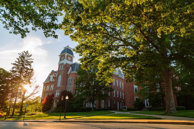 Thick green trees stand in front of Miller and Hanna Halls as the sun's rays peak through the leaves.