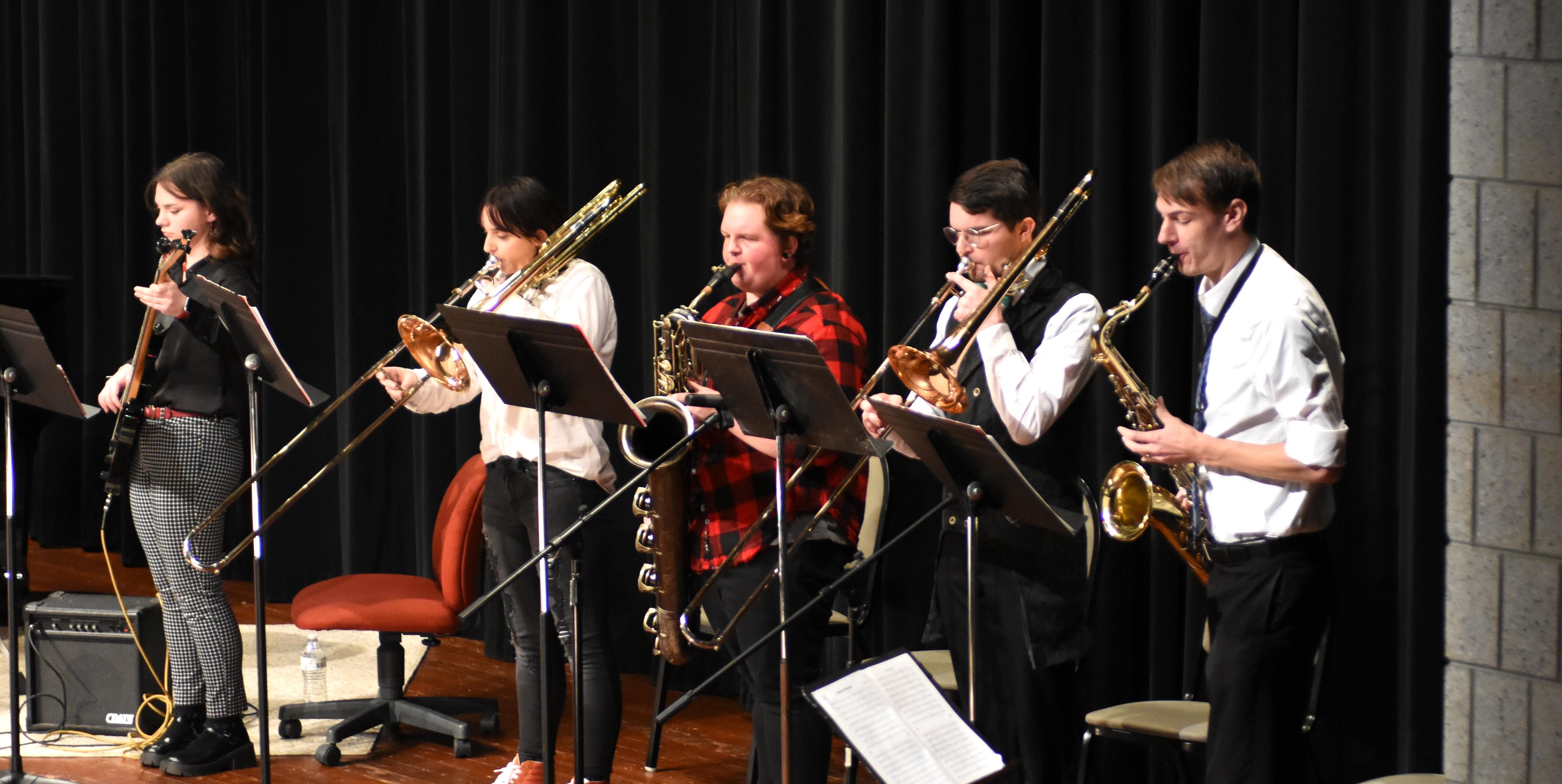 Image of five students playing their respective instruments in the fall 2023 jazz ensemble concert. The students stand on the Goodwin Performing Arts Center stage which has a brown wooden floor and a black curtain.