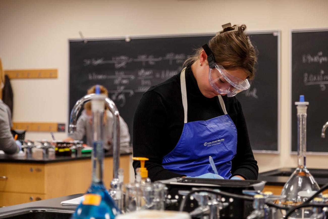 Waynesburg student works in the science lab with an apron and goggles