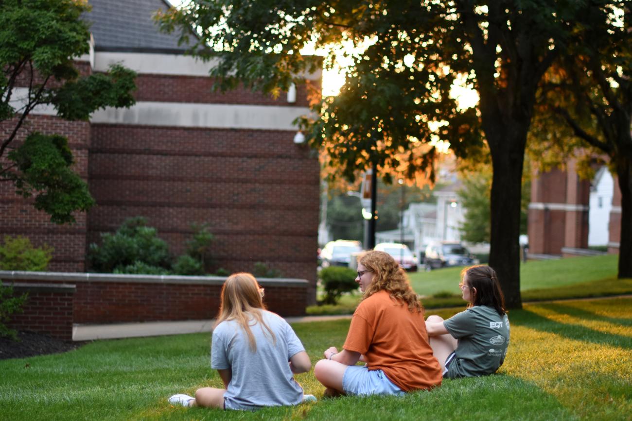 Three students sitting outside in the grass on a summer day