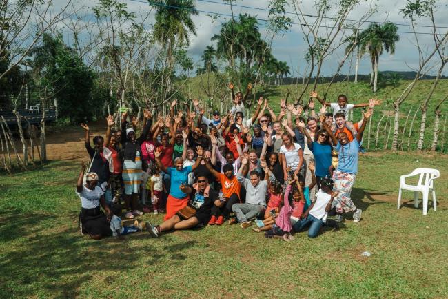 Waynesburg trip participants gather for a group photo with the women and children of Luma Largo. The group holds their hands in the air and smiles for the camera. 